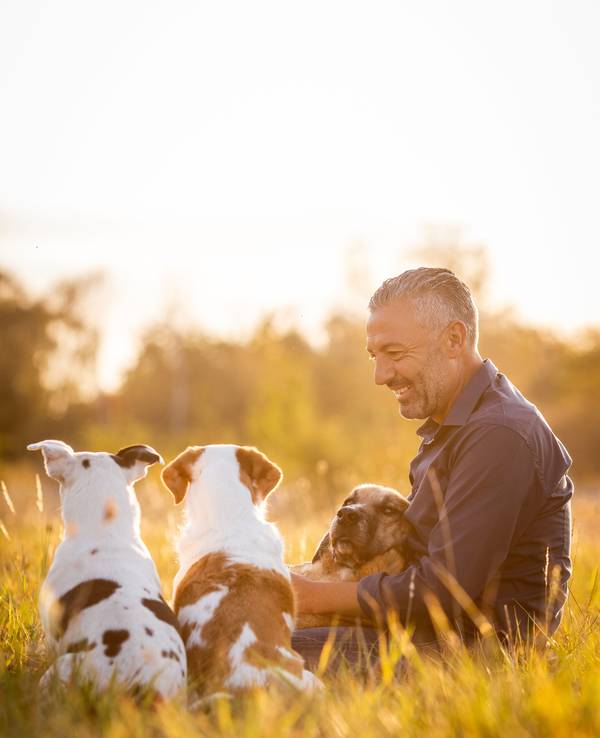 Ganz entspannt sitzt Hund-Mensch-Coach Andreas Ohligschläger mit drei Hunden in einer Wiese und lächelt.