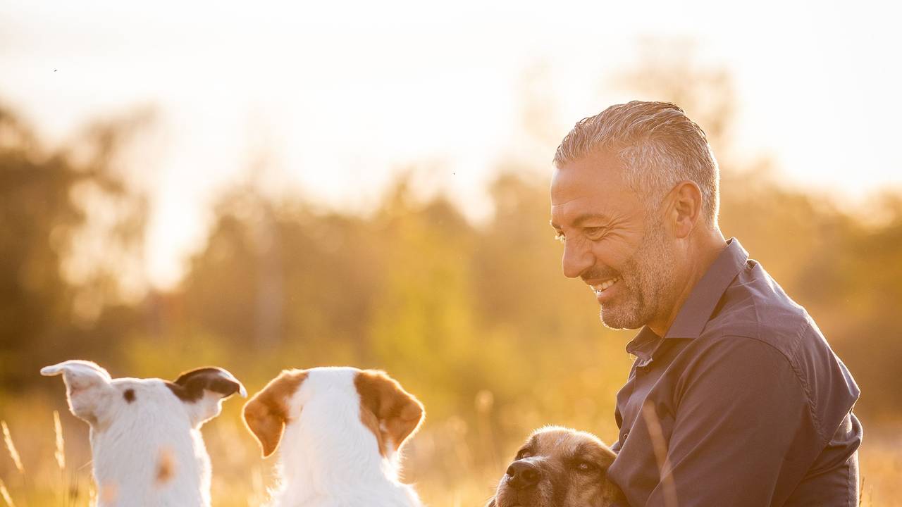 Ganz entspannt sitzt Hund-Mensch-Coach Andreas Ohligschläger mit drei Hunden in einer Wiese und lächelt.