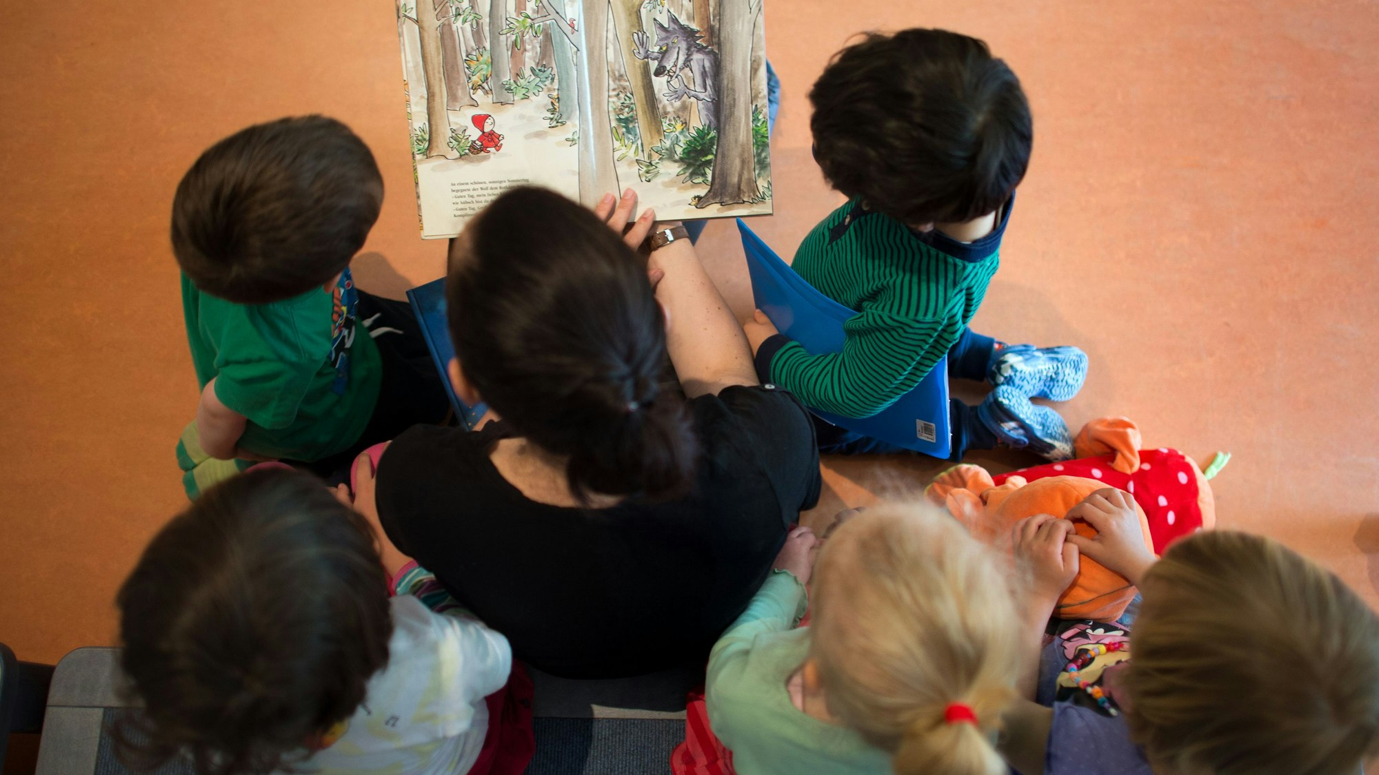 Eine Frau sitzt auf einem Stuhl und liest fünf Kindern etwas vor. Die sechs Personen sind von oben zu sehen.