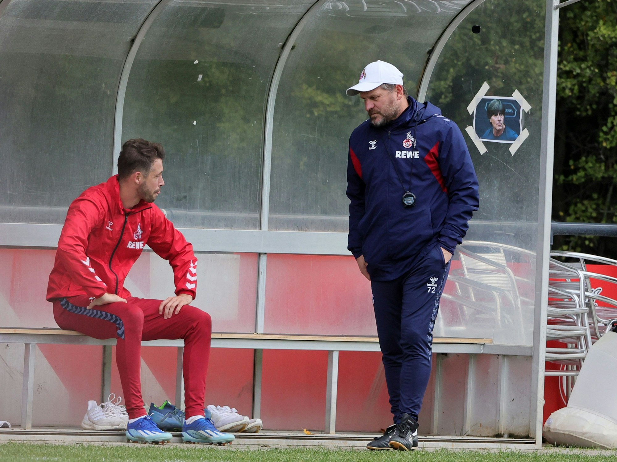 Steffen Baumgart spricht mit Mark Uth, im Hintergrund hängt ein Bild von Jogi Löw.