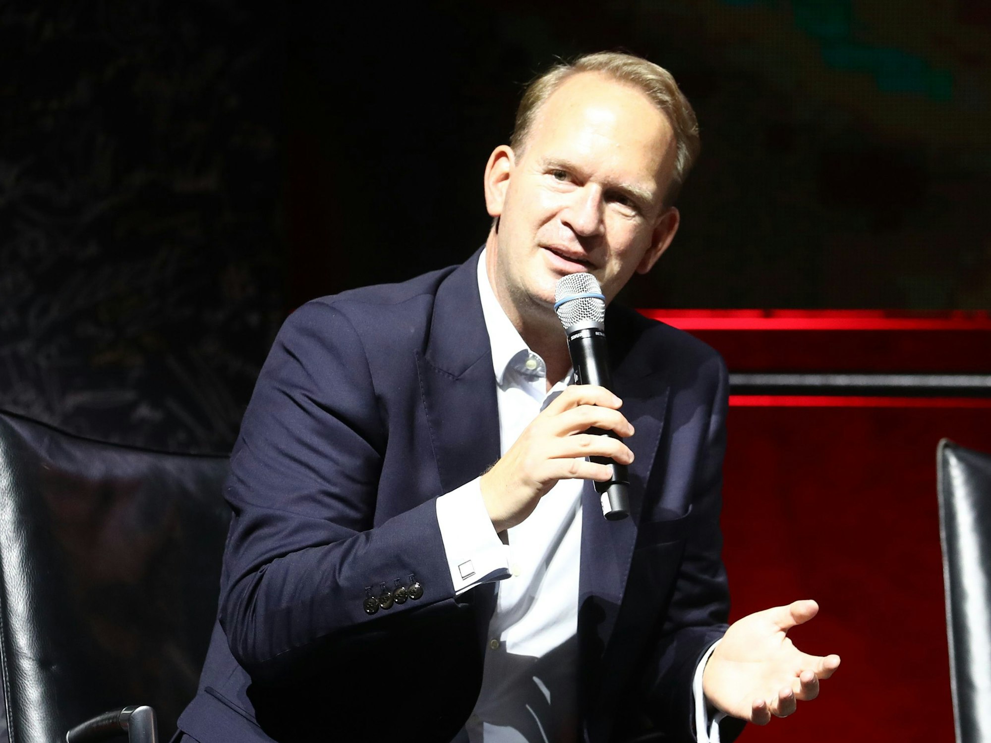 Stefan Löcher mit Mikrofon auf einer Pressekonferenz. Er trägt einen dunklen Anzug.
