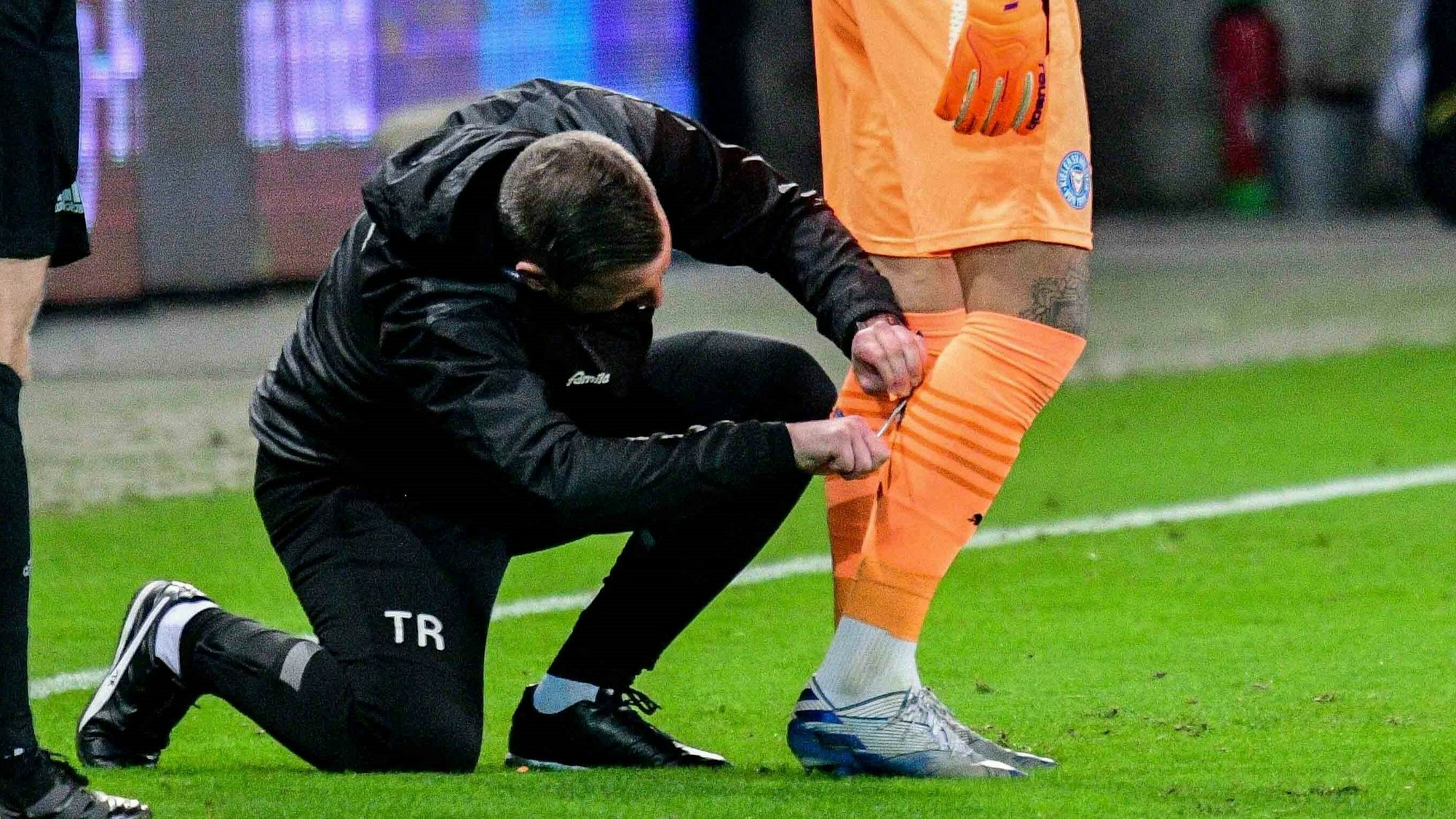 Ein Betreuer schnibbelt an den Stutzen von Holstein Kiels Torwart Tim Schreiber.