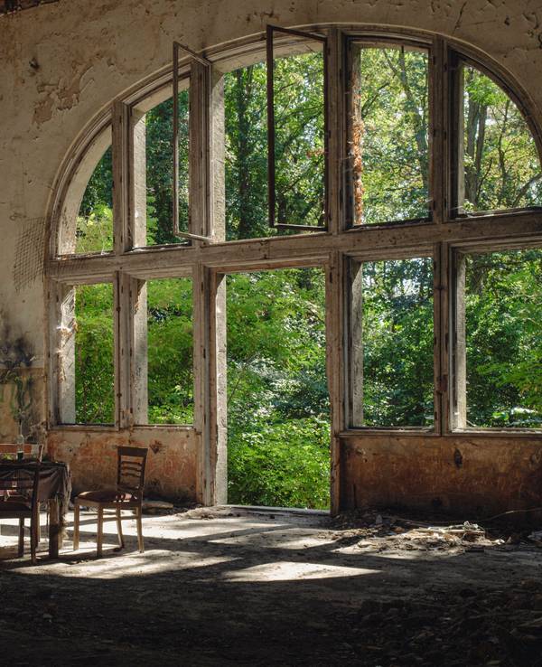Symbolbild Beelitz Heilstätten Deutschland, Beelitz am 09.09.2023: In der Ruine des Speisesaals im Alpen-Haus stehen fünf alte Stühle und um einen alten Tisch mit Gläsern und eine Flasche vor einem riesigen Fenster.