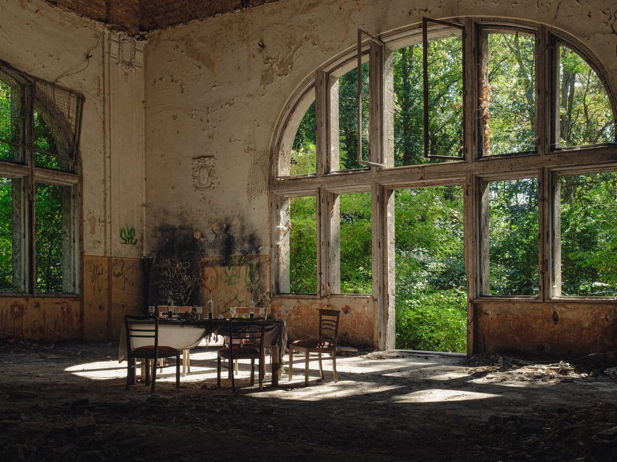 Symbolbild Beelitz Heilstätten Deutschland, Beelitz am 09.09.2023: In der Ruine des Speisesaals im Alpen-Haus stehen fünf alte Stühle und um einen alten Tisch mit Gläsern und eine Flasche vor einem riesigen Fenster.