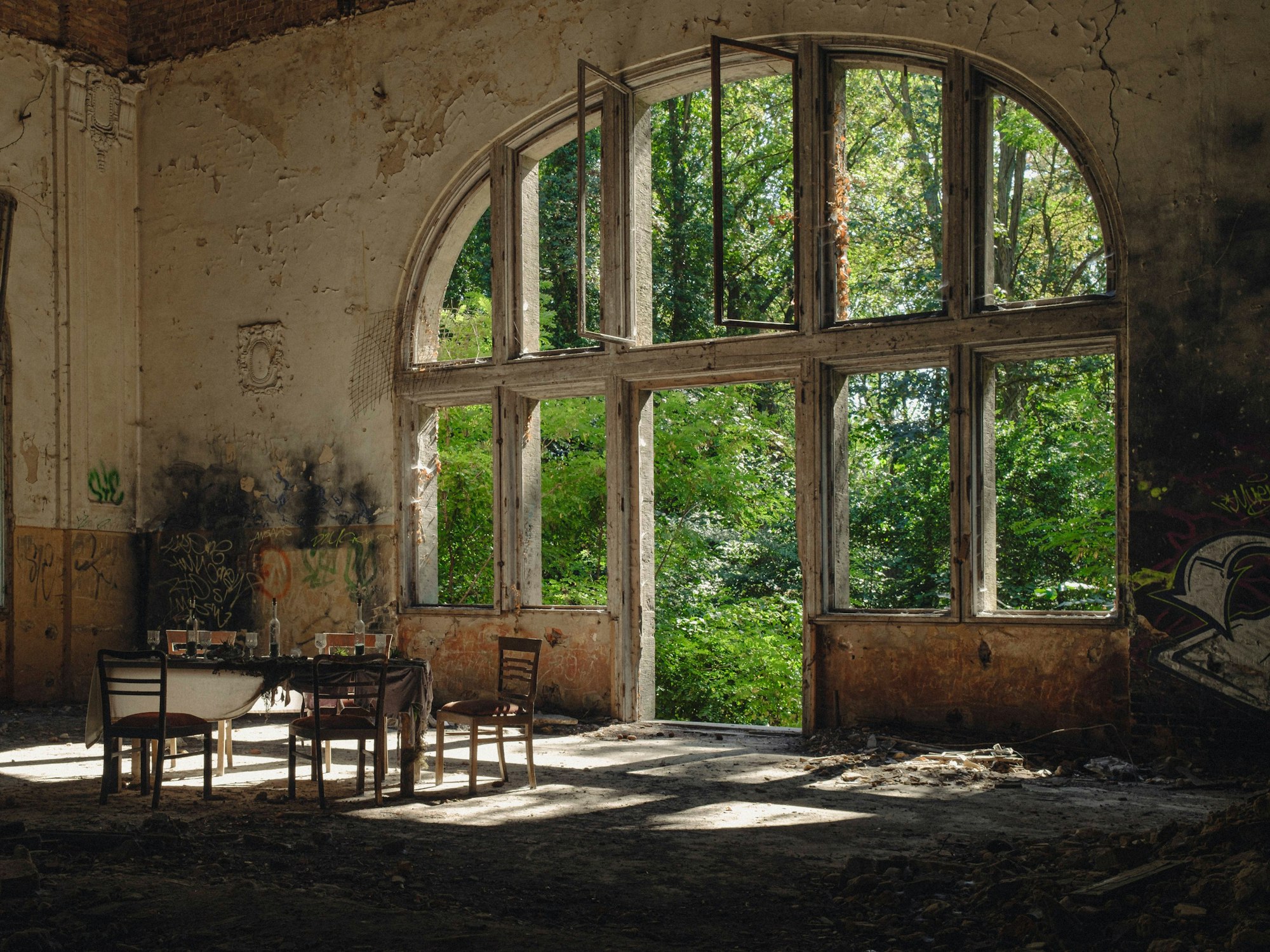 Symbolbild Beelitz Heilstätten Deutschland, Beelitz am 09.09.2023: In der Ruine des Speisesaals im Alpen-Haus stehen fünf alte Stühle und um einen alten Tisch mit Gläsern und eine Flasche vor einem riesigen Fenster.