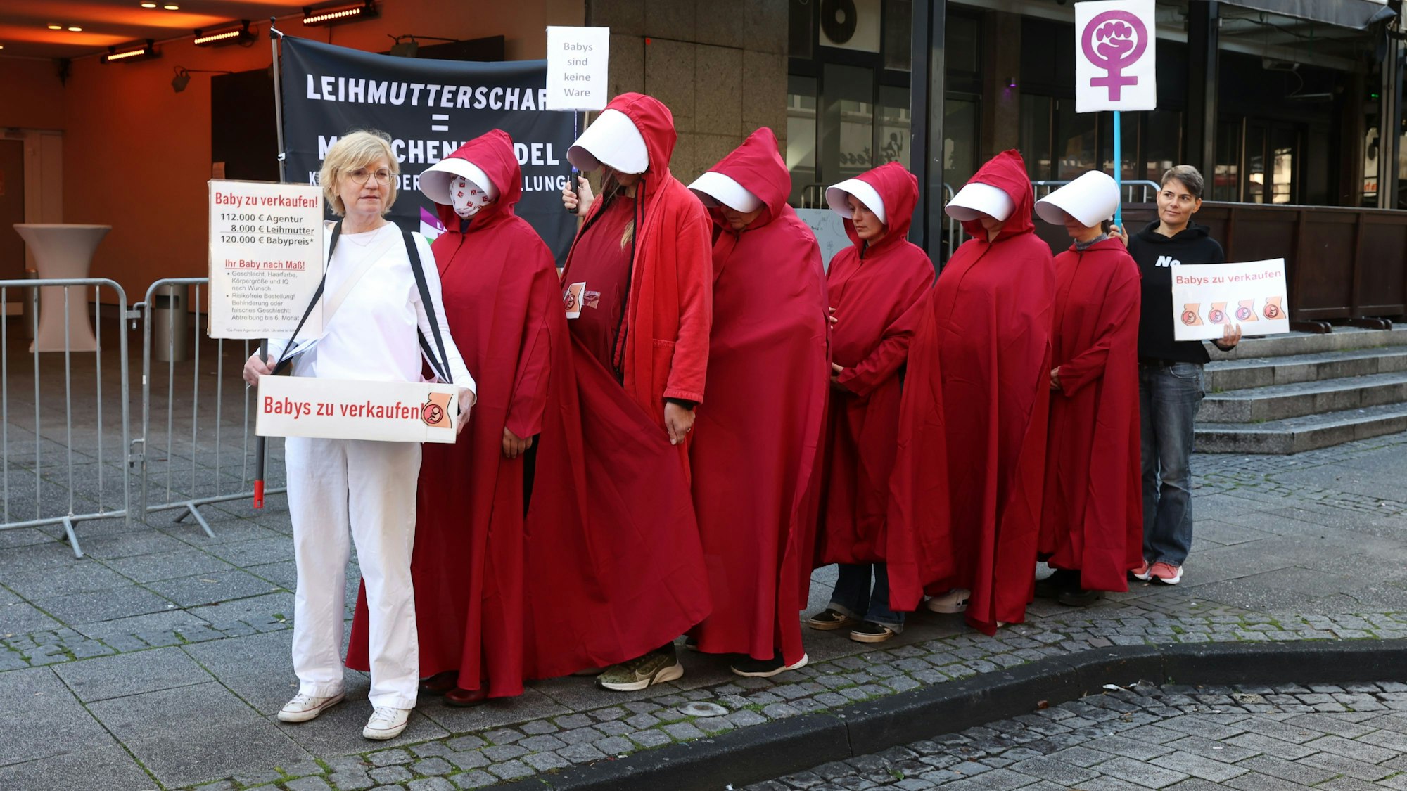 Mehrere Frauen stehen vor einer Messehalle und protestieren mit Schildern in der Hand. Einige von ihnen sind komplett in rote Roben gehüllt.