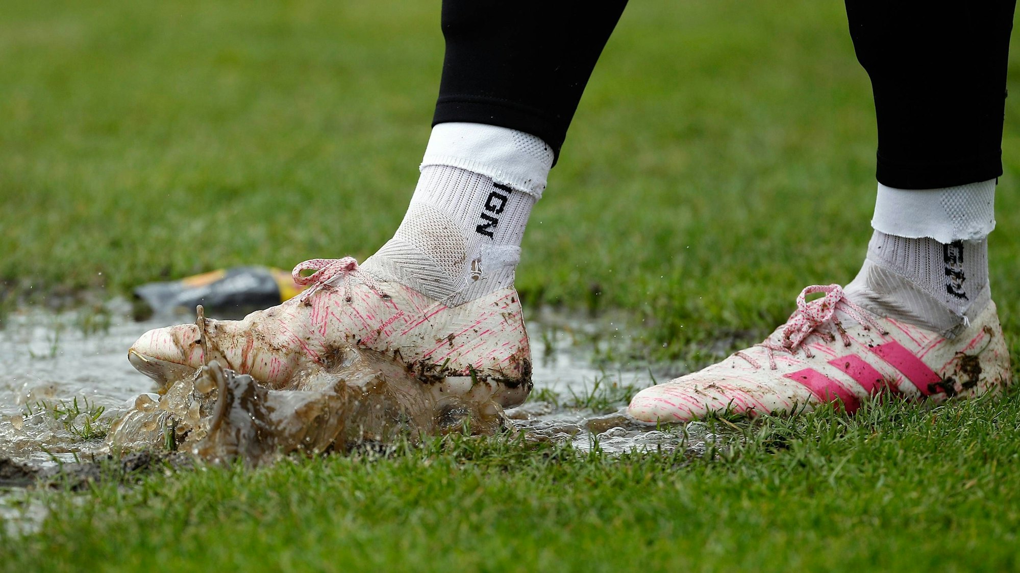 Ein Fußballer steht auf dem Rasen in einer Pfütze, auf dem Bildausschnitt sind nur seine Füße zu sehen.