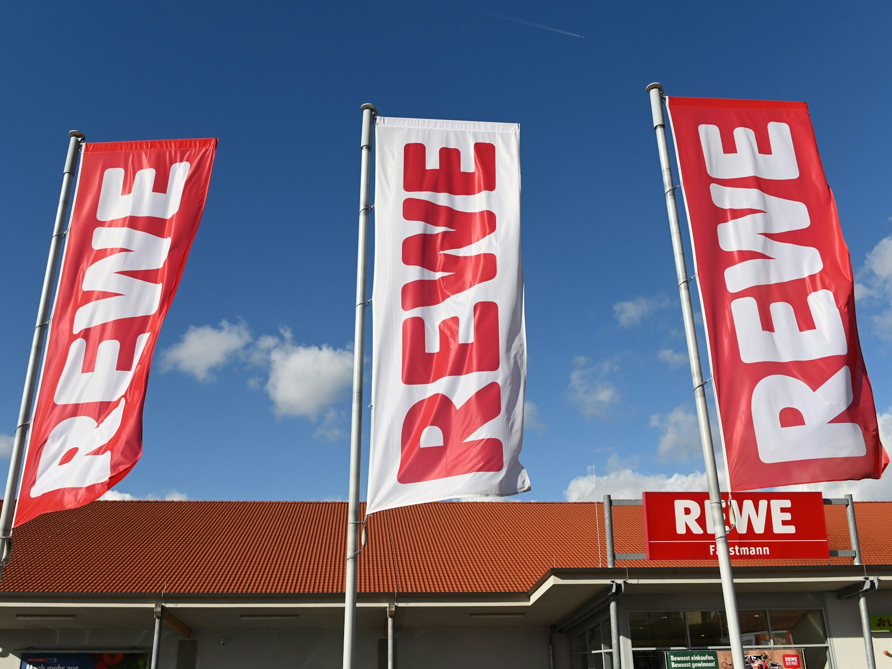 Fahnen mit dem Logo des Lebensmitteleinzelhändlers Rewe wehen vor einer Filiale.