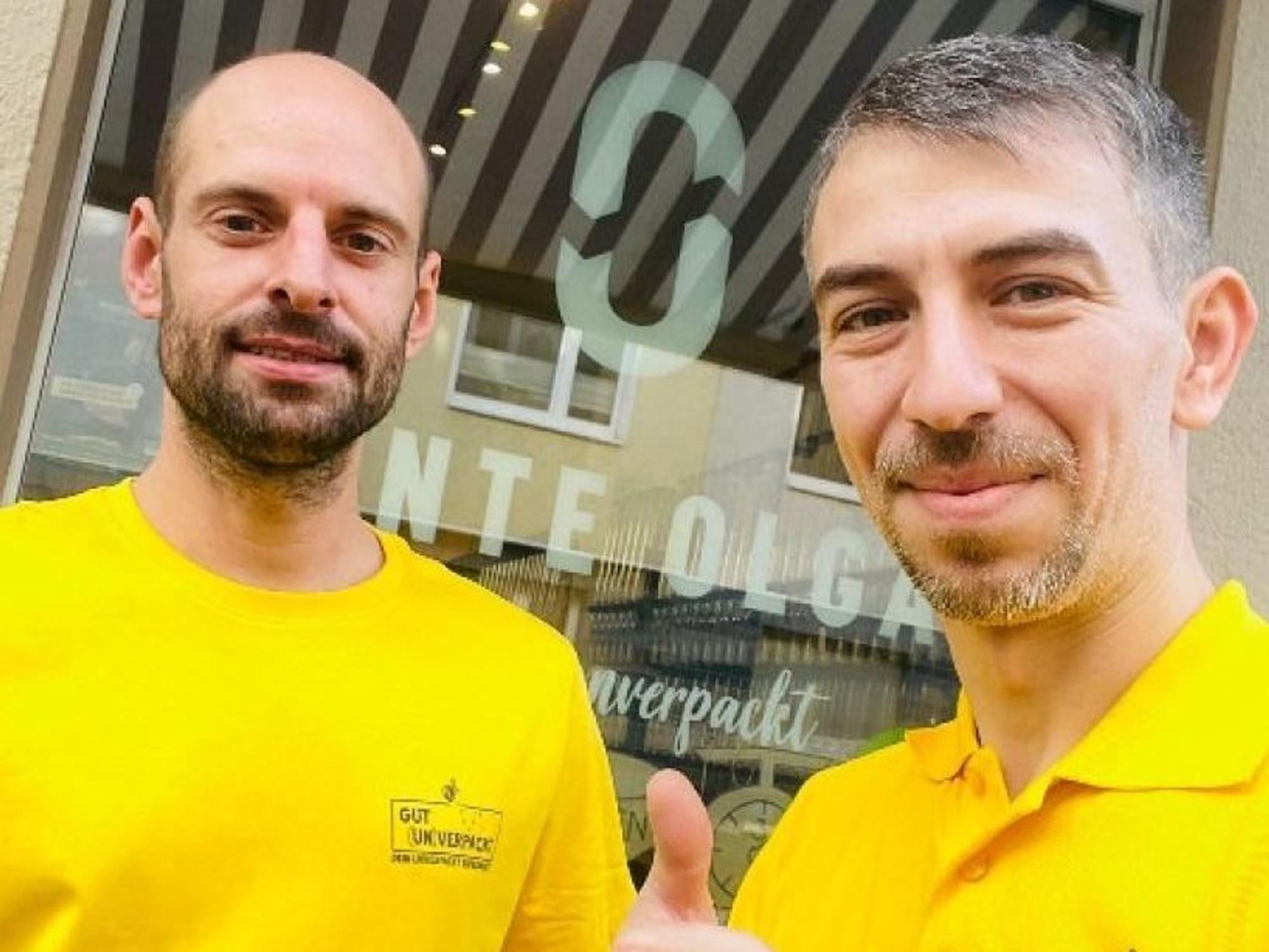 Zwei Männer in gelben Poloshirts stehen vor einem Schaufenster und schauen in die Kamera.