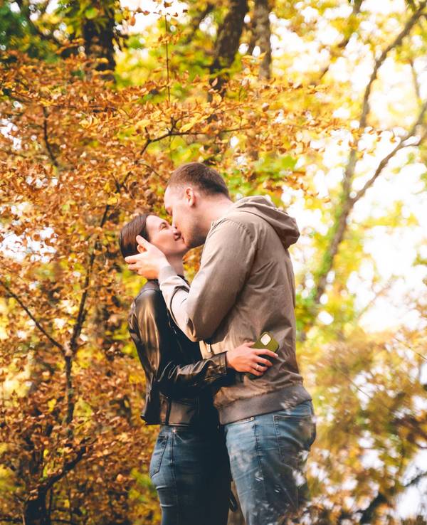 A young Caucasian couple is kissing in the Autumn forest, embracing each other at sunset. Weekend time with family and beautiful romantic love story. Space for text copy