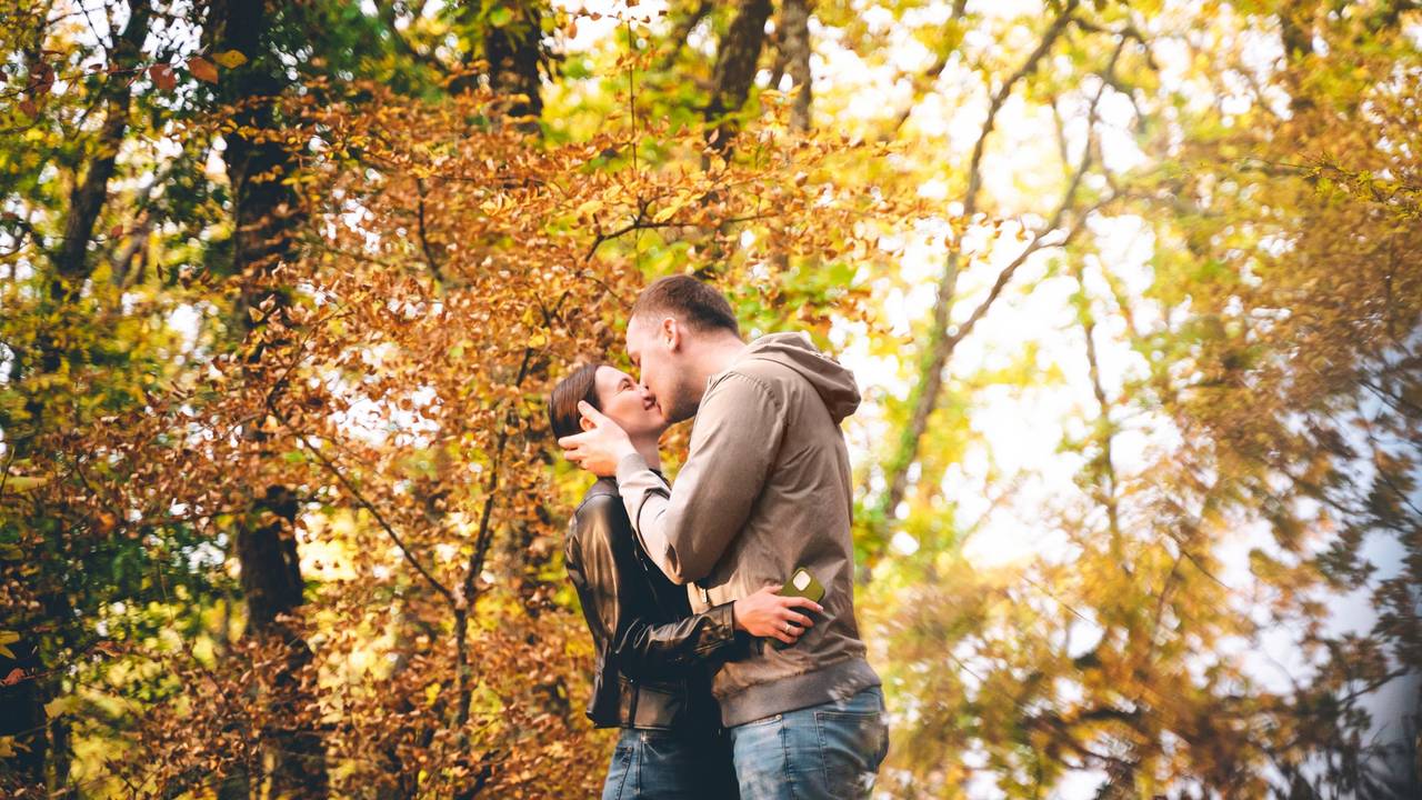 A young Caucasian couple is kissing in the Autumn forest, embracing each other at sunset. Weekend time with family and beautiful romantic love story. Space for text copy