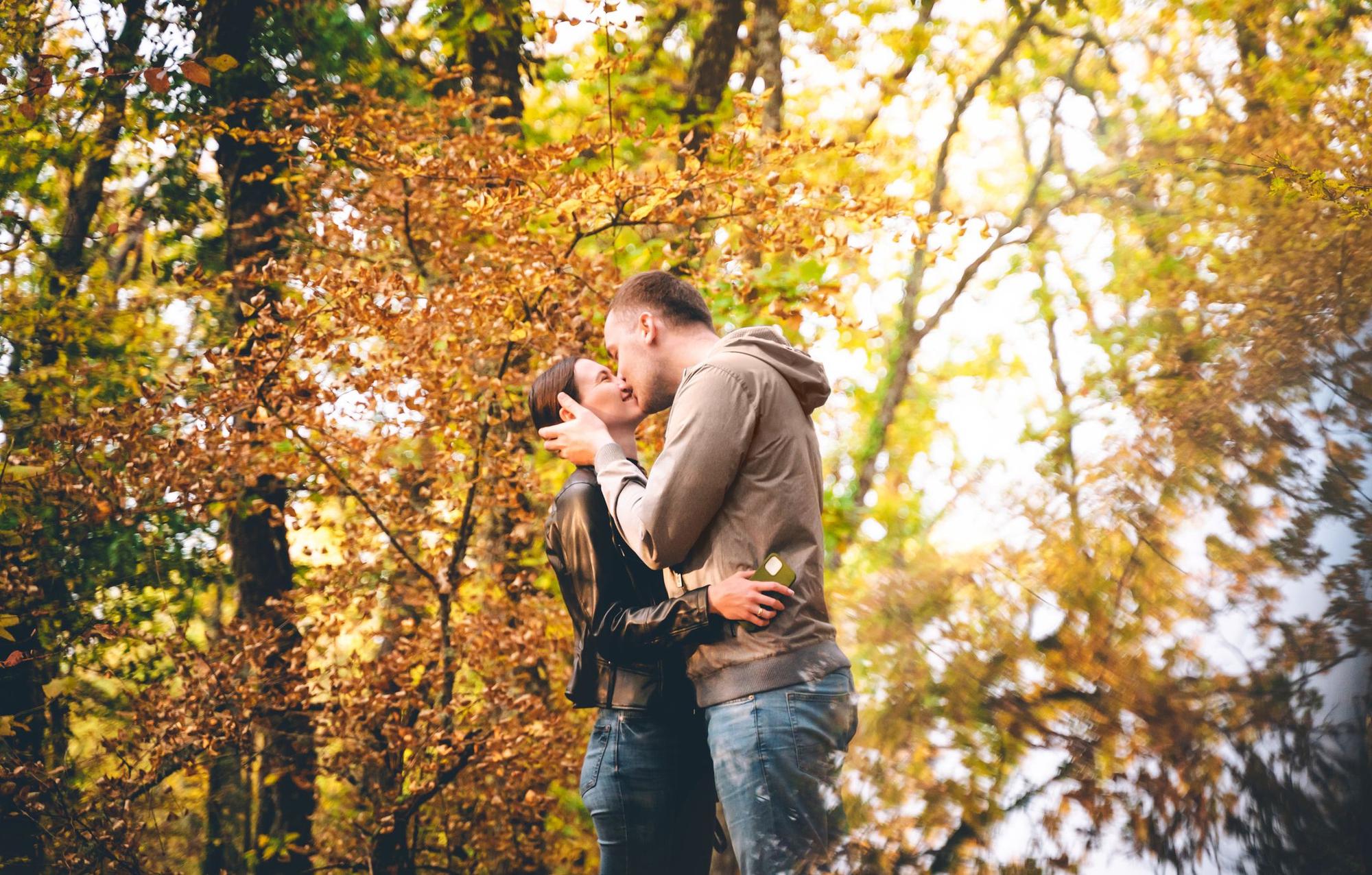 A young Caucasian couple is kissing in the Autumn forest, embracing each other at sunset. Weekend time with family and beautiful romantic love story. Space for text copy