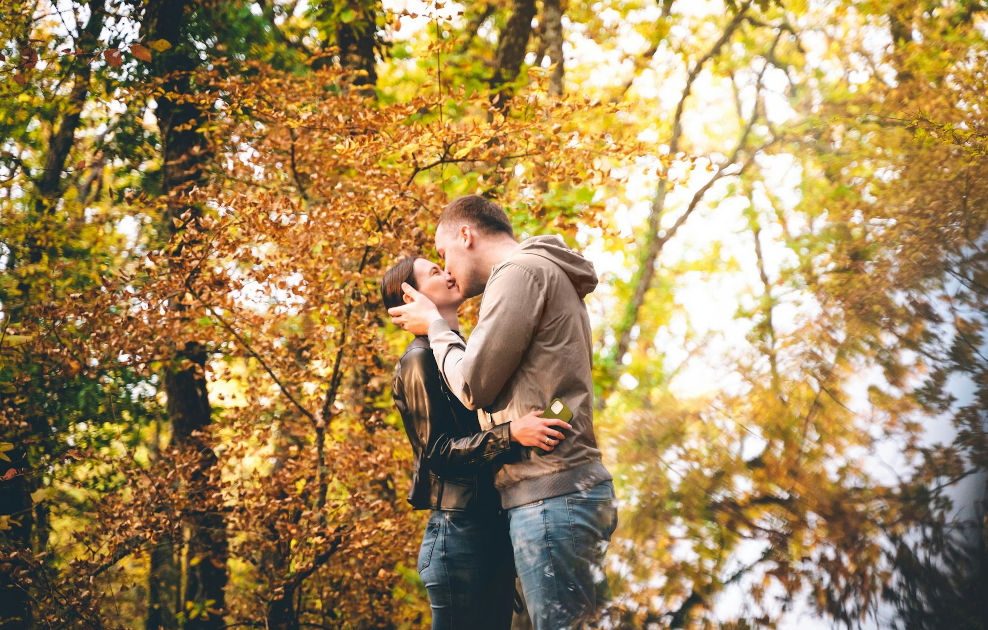 A young Caucasian couple is kissing in the Autumn forest, embracing each other at sunset. Weekend time with family and beautiful romantic love story. Space for text copy