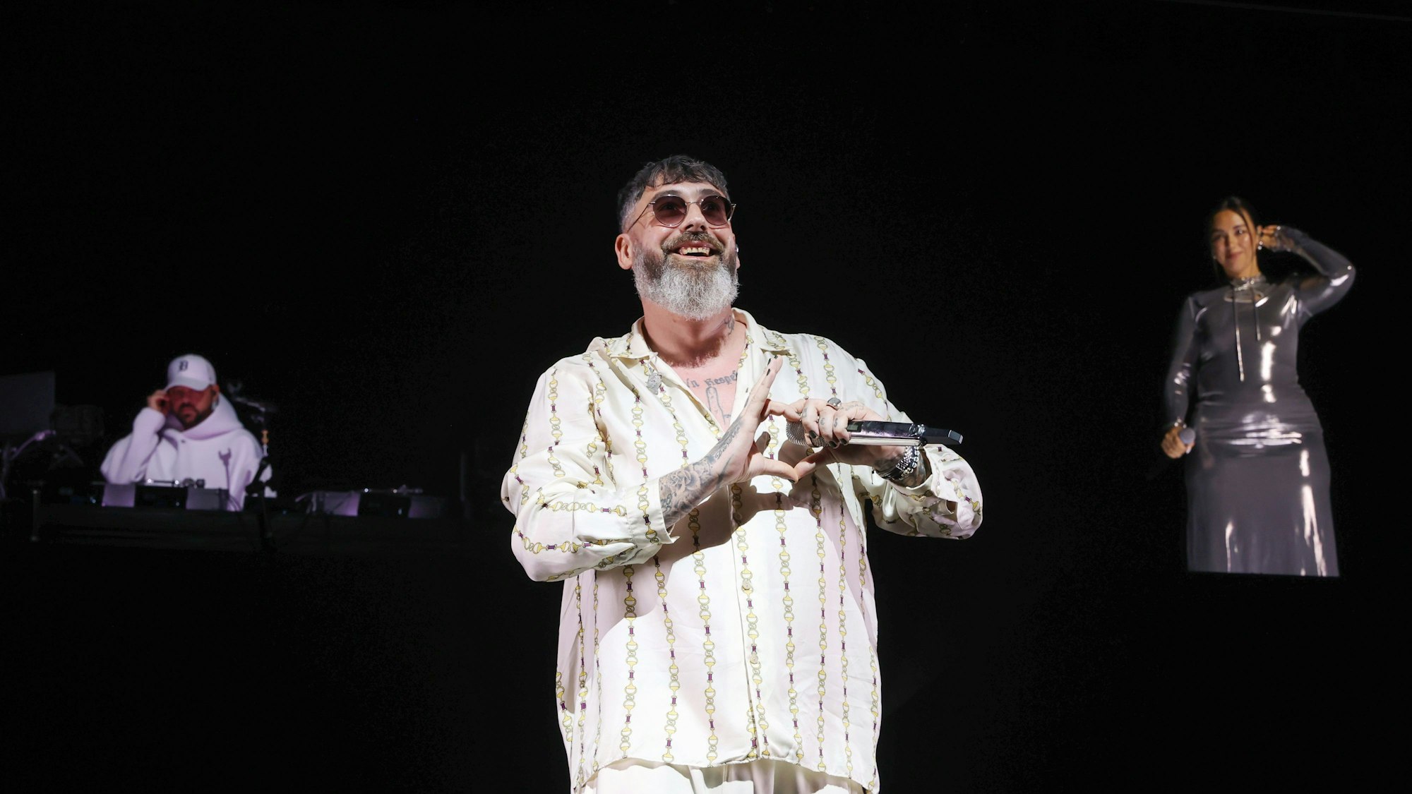 Sido zeigt mit seinen Fingern ein Herz und lächelt. Im Hintergrund ein DJ und die Background-Sängerin.