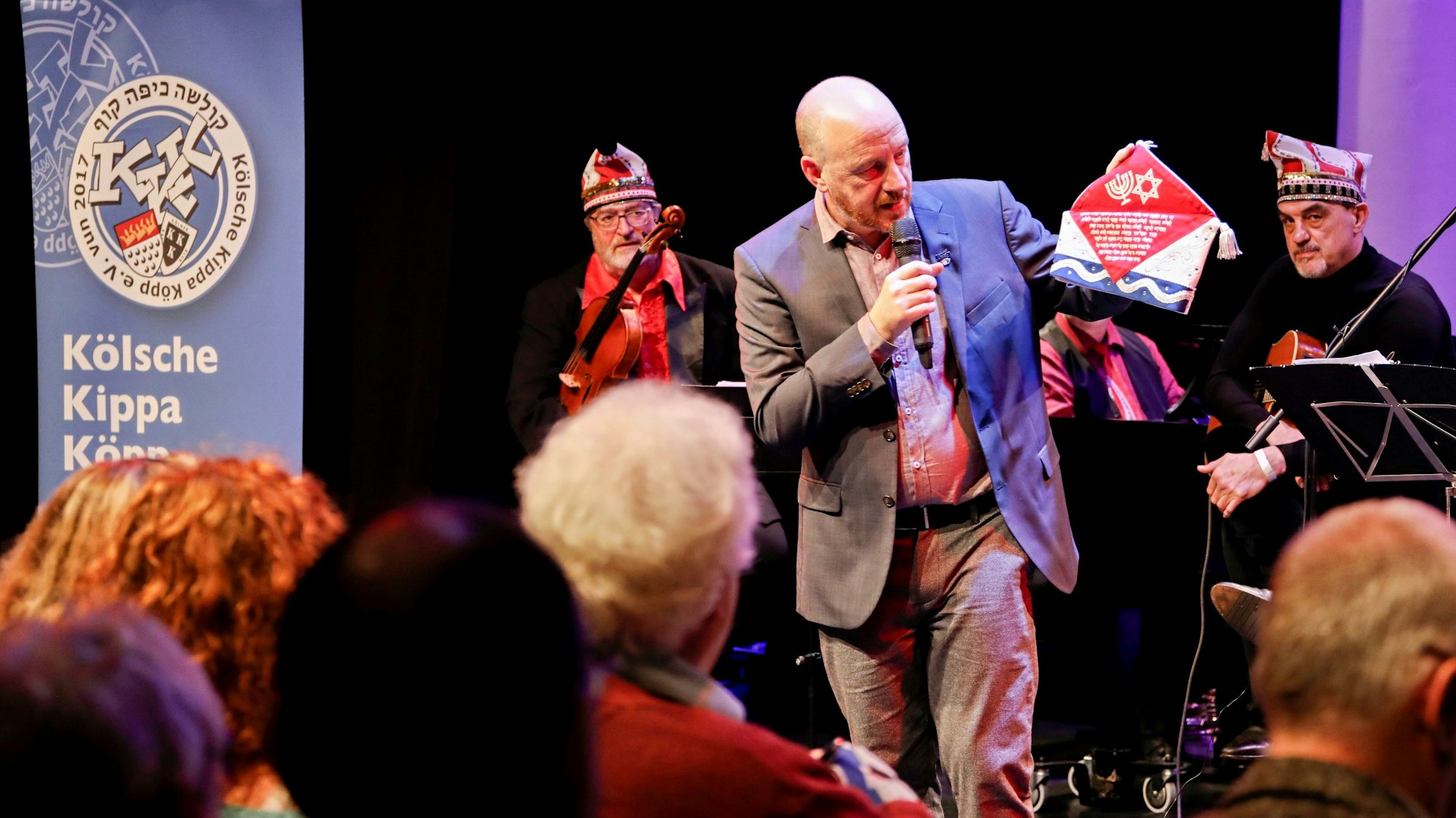 Ein Mann im Anzug steht auf einer Bühne und zeigt dem Publikum eine Karnevalsmütze.