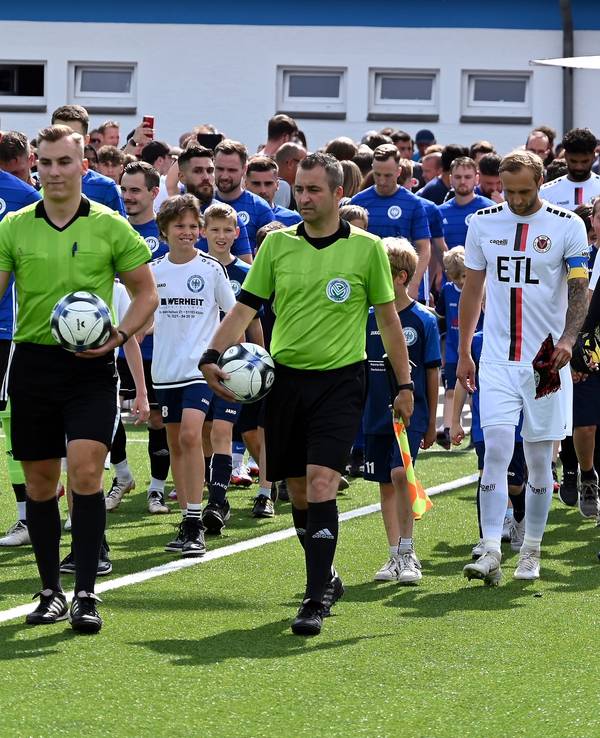 Zwei Fußball-Mannschaften in blauen und weißen Trikots laufen gemeinsam mit den Schiedsrichtern auf das Feld.