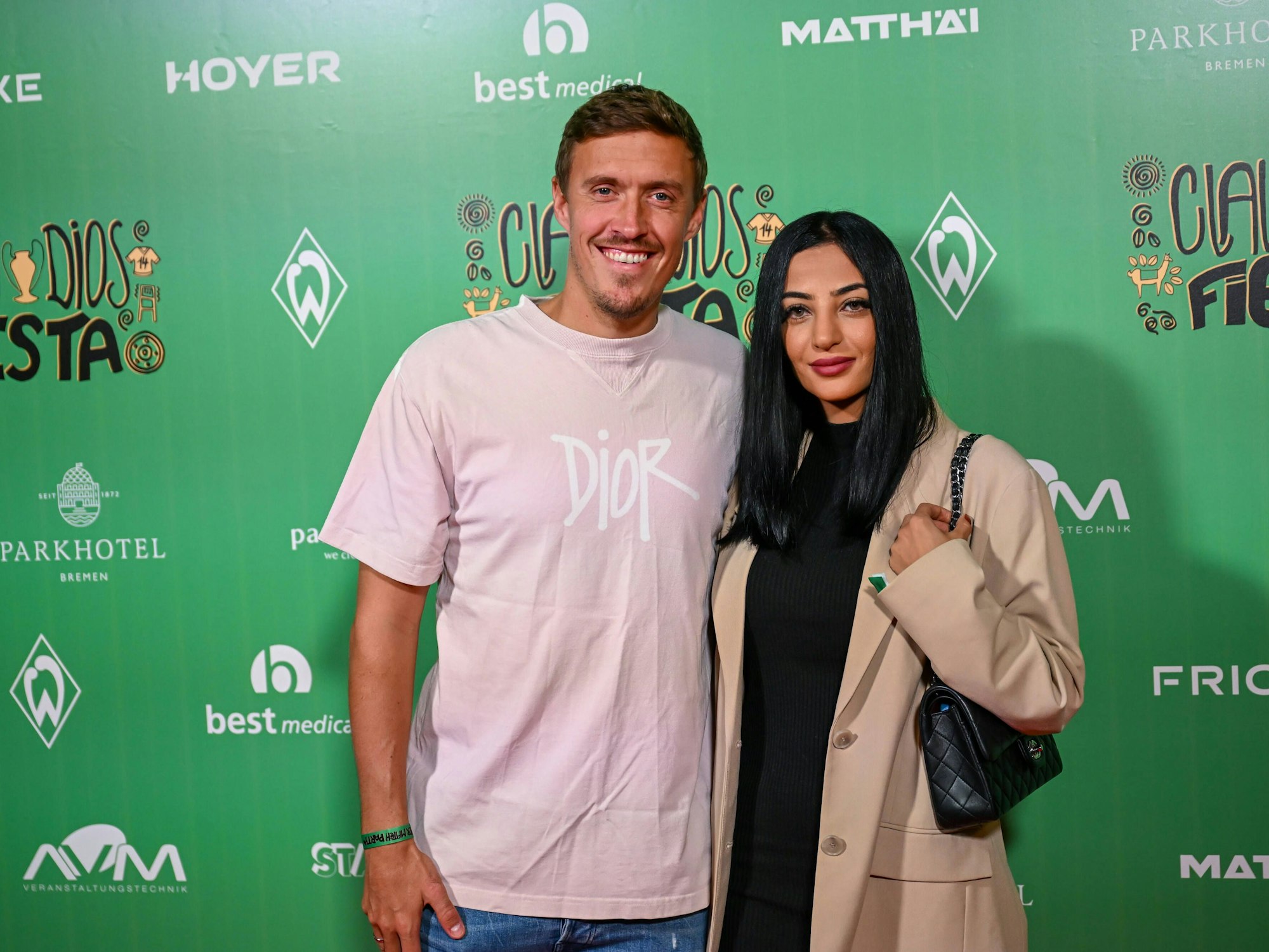 Das Foto zeigt Dilara und Max Kruse im Wohninvest Weserstadion in Bremen vor einer Wand, auf welcher Logos verschiedener Sponsoren des SV Werder Bremen abgedruckt sind.