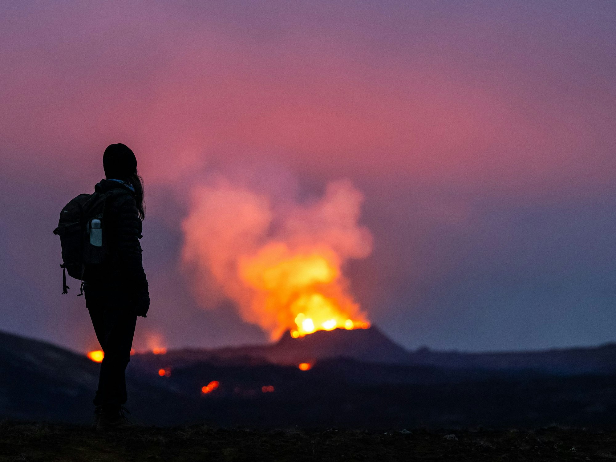 Lava tritt aus dem Krater eines Vulkans in der Nähe des Berges Litli-Hrútur aus, etwa 40 Kilometer südwestlich von Reykjavik. Im Vordergrund steht eine junge Frau und blickt auf die Flammen, hier im Juli 2023.