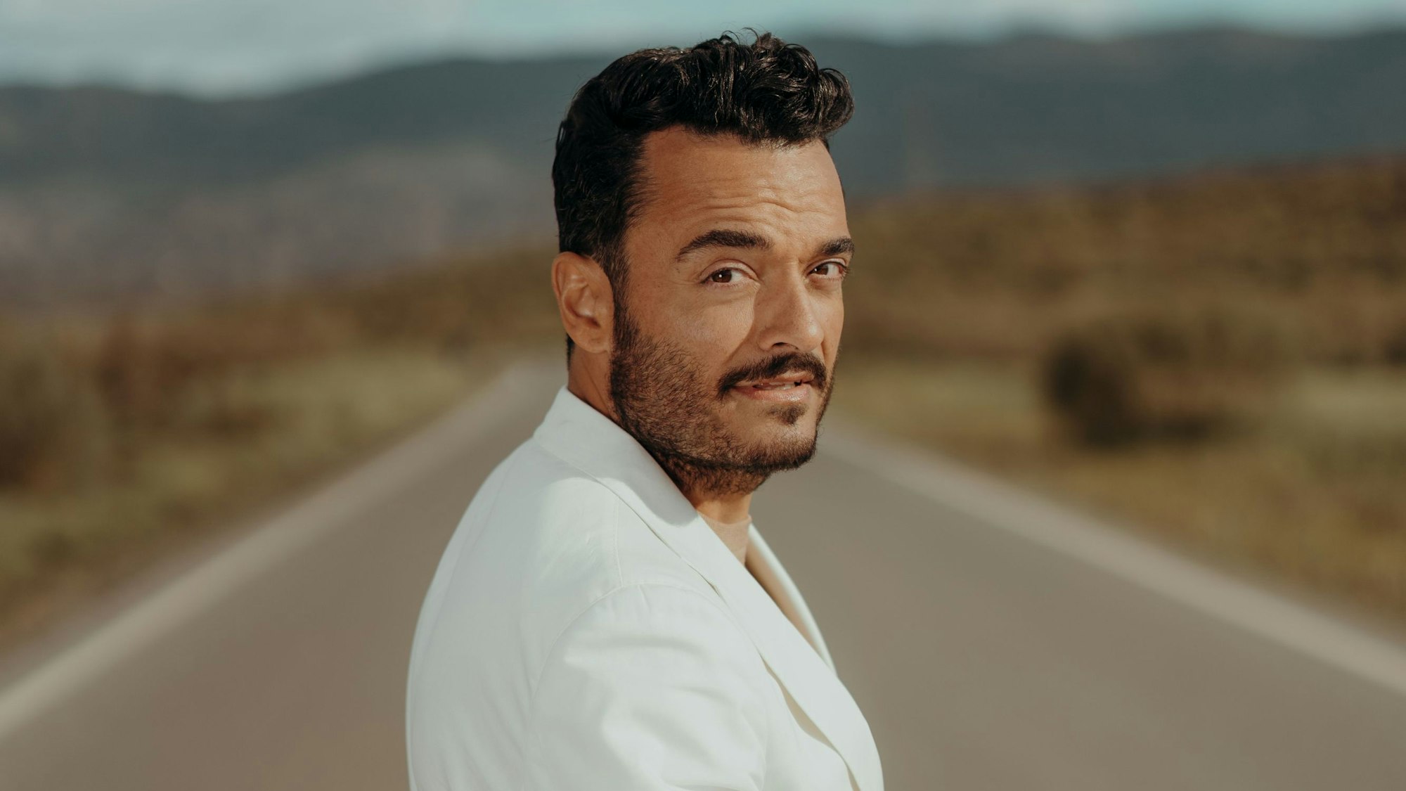 Giovanni Zarrella trägt einen weißen Anzug und blickt auf einer Straße über die Schulter in die Kamera.