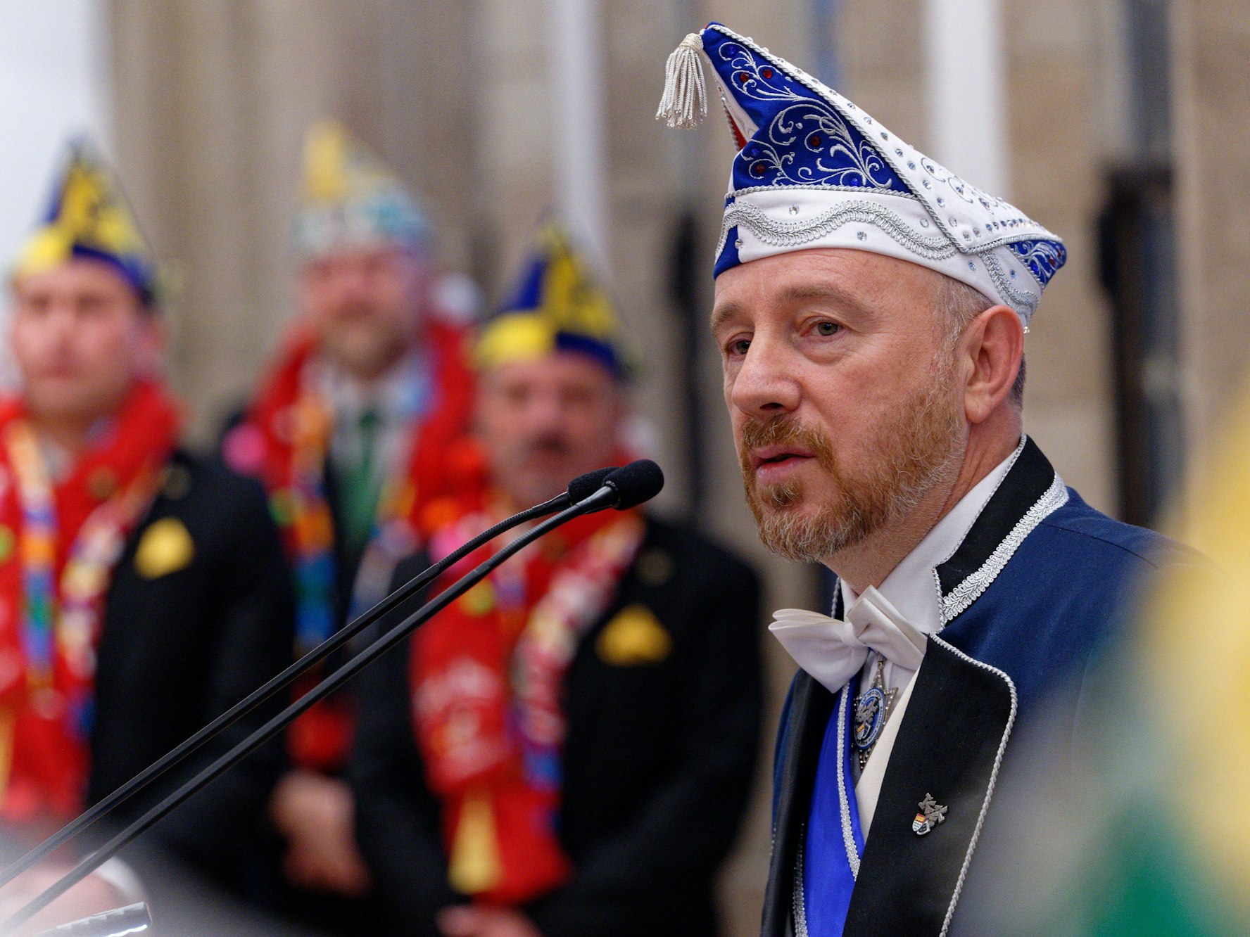 Aaron Knappstein mit Karnevalsmütze bei seiner Rede, vor ihm Mikrofone.