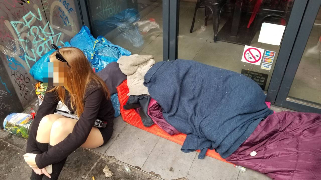 Ein Mädchen sitzt am 11.11.2023 in einem Eingang zu einer Gastro auf der Zülpicher Straße – hinter ihr liegt ein schlafender Obdachloser.