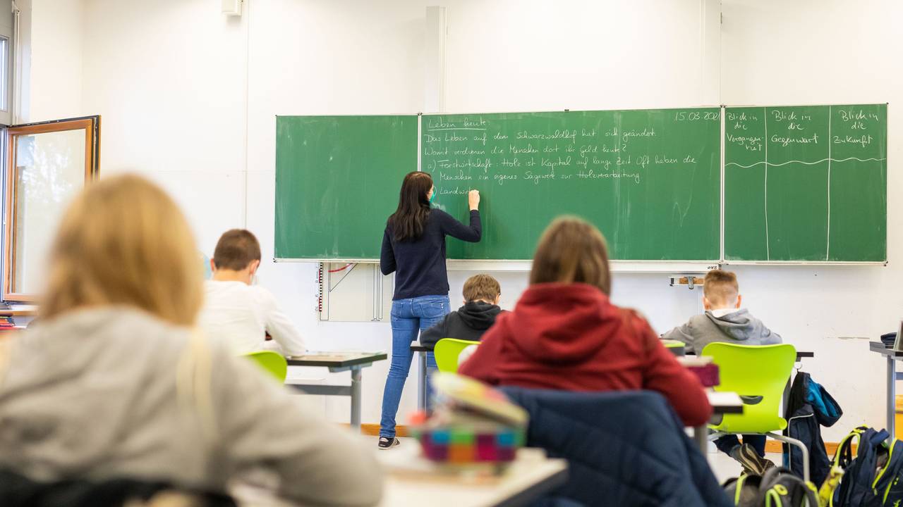 Eine Lehrerin schreibt etwas an die Tafel, die Schulkinder schreiben mit.