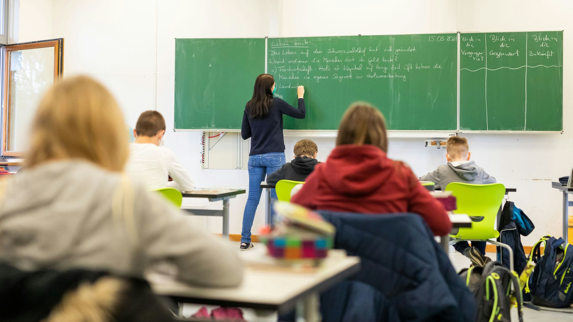 Eine Lehrerin schreibt etwas an die Tafel, die Schulkinder schreiben mit.