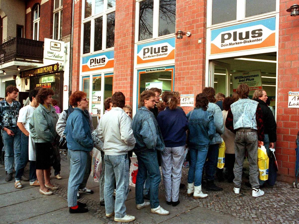 Das Archivbild vom 11.11.1989 zeigt eine lange Schlange von DDR-Bürgerinnen und -Bürgern, die nach der Maueröffnung zu Tausenden nach West-Berlin und die Bundesrepublik strömten. Und damit auch in die Supermärkte, wie Plus. Heute ist Plus längst verschwunden.
