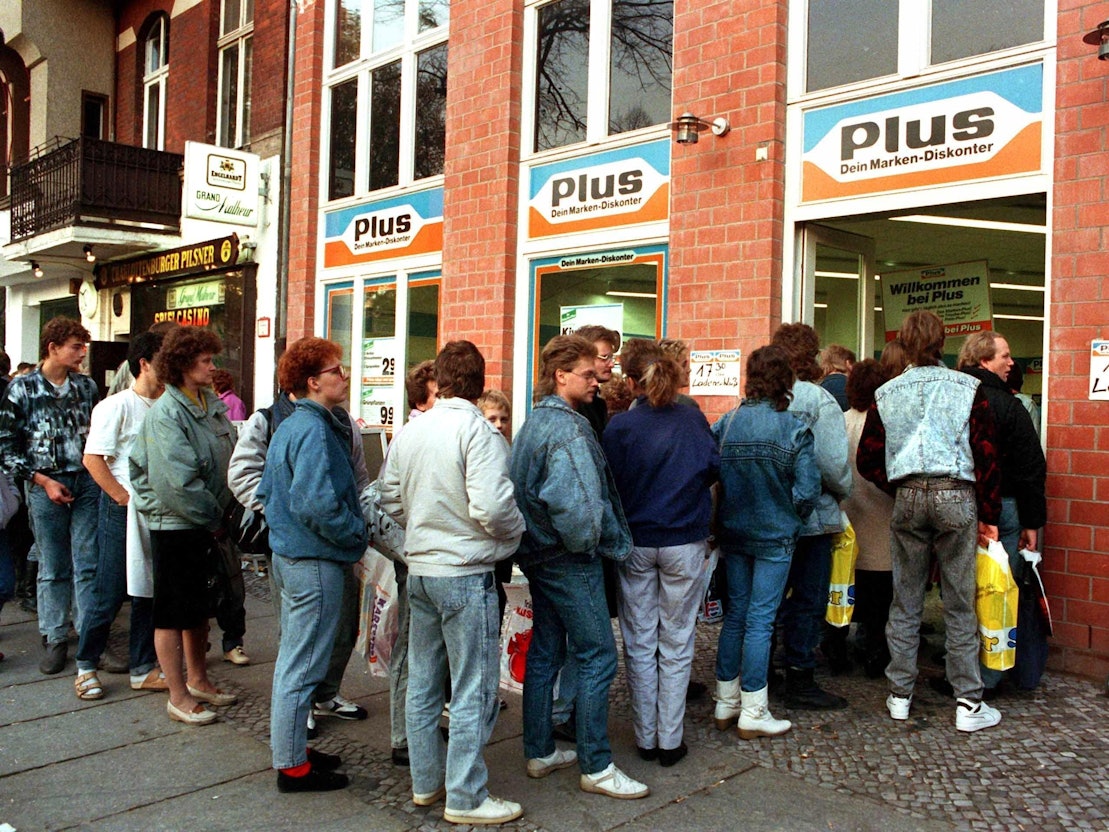 Das Archivbild vom 11.11.1989 zeigt eine lange Schlange von DDR-Bürgerinnen und -Bürgern, die nach der Maueröffnung zu Tausenden nach West-Berlin und die Bundesrepublik strömten. Und damit auch in die Supermärkte, wie Plus. Heute ist Plus längst verschwunden.