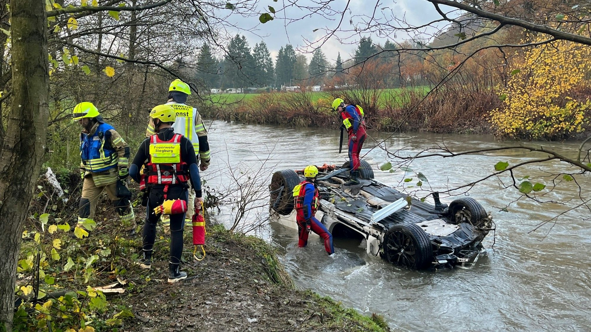 Ein Auto liegt auf dem Dach in einem Fluss, Rettungskräfte stehen auf und an dem Wagen sowie am Ufer.