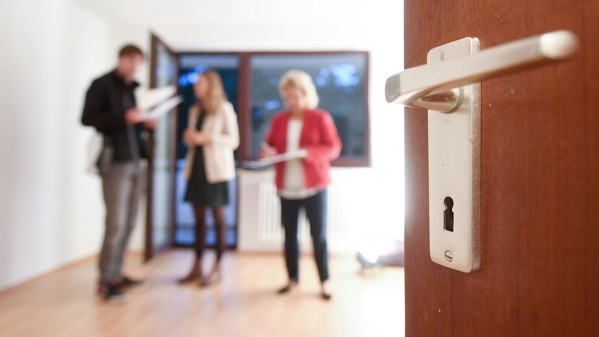 Eine Maklerin spricht mit Interessenten bei einer Wohnungsbesichtigung, während im Vordergrund eine Türklinke zu sehen ist.