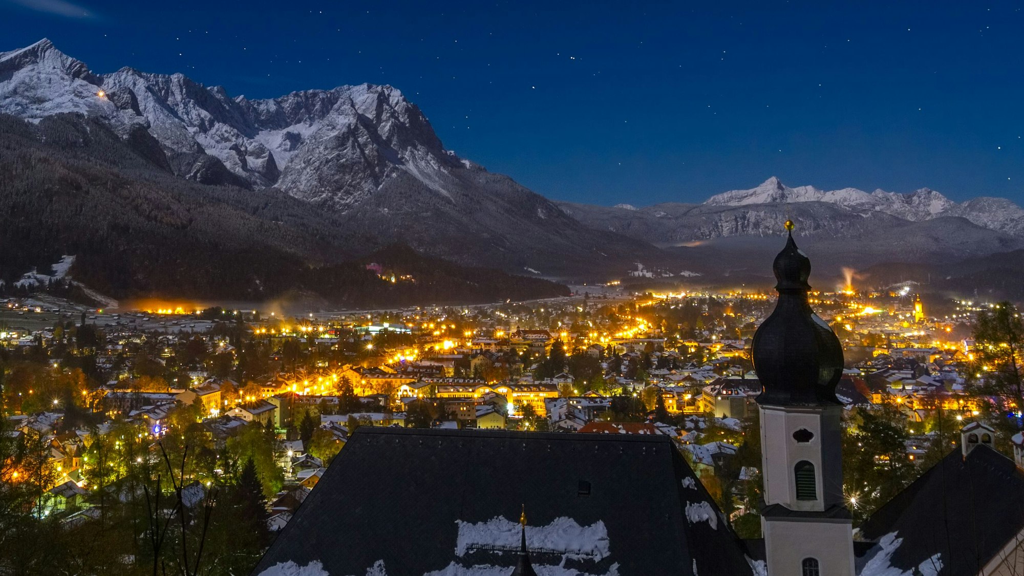 Blick auf das bayerische Garmisch-Partenkirchen bei Nacht. Die Stadt ist umrahmt von den schneebedeckten Alpen.