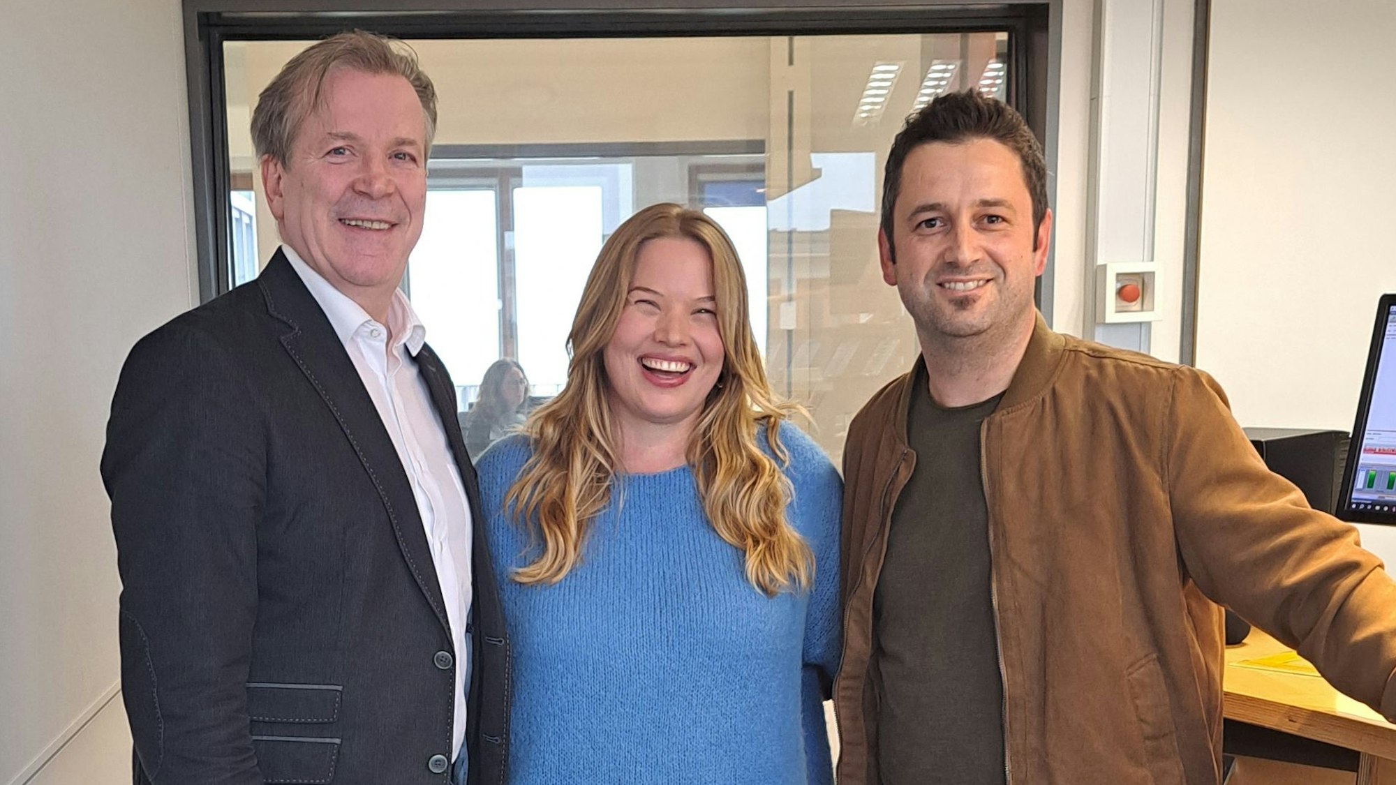 Zwei Männer und eine Frau stehen in einem Tonstudio und lächeln in die Kamera.