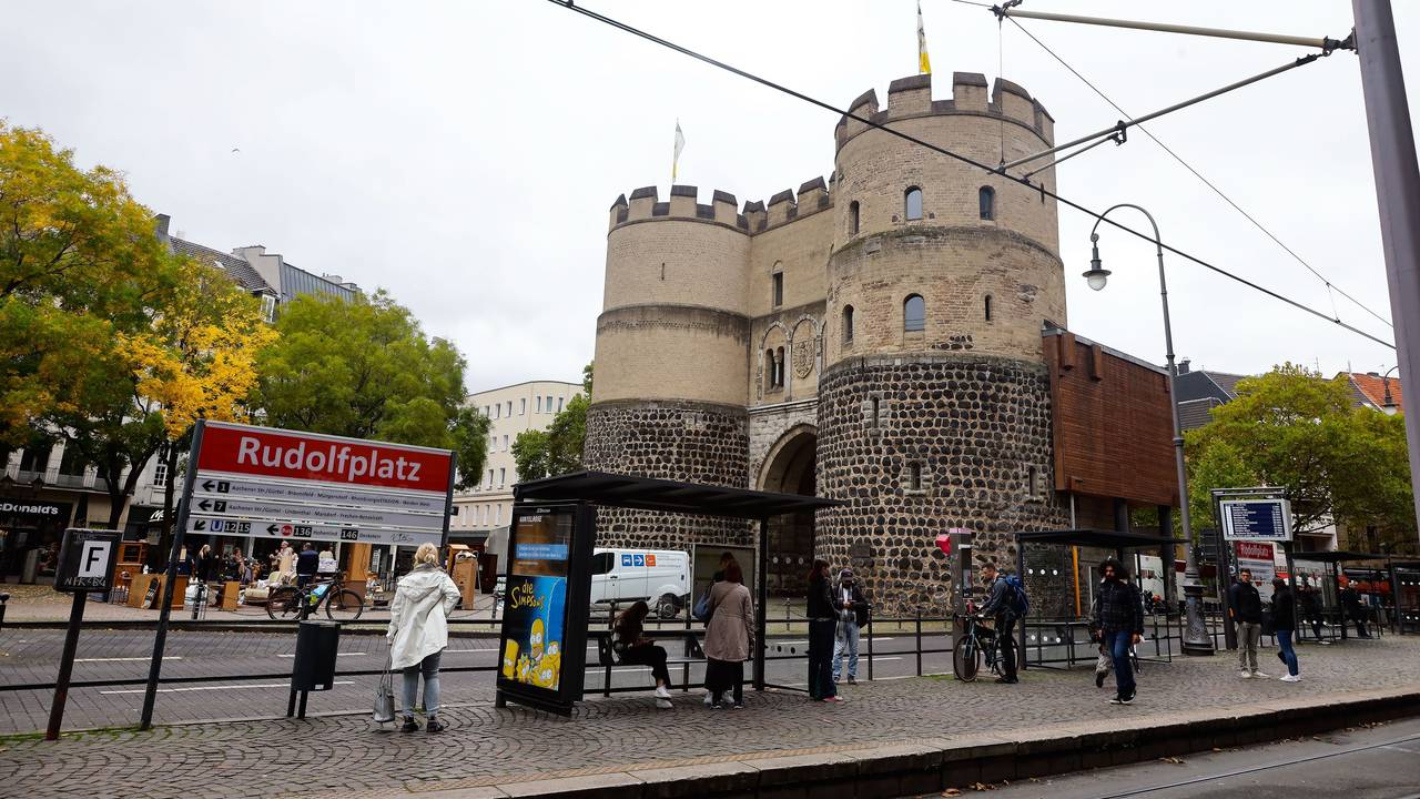An einer Haltestelle mit der Aufschrift Rudolfplatz stehen mehrere Menschen.