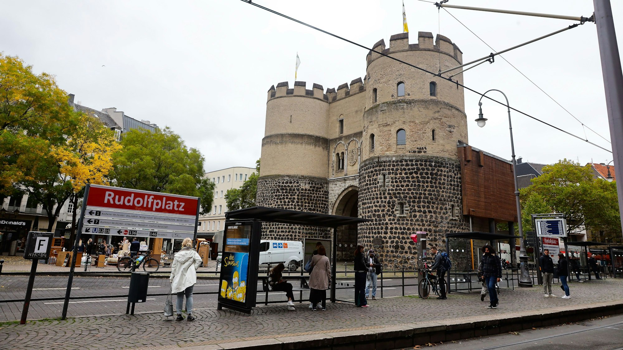 An einer Haltestelle mit der Aufschrift Rudolfplatz stehen mehrere Menschen.