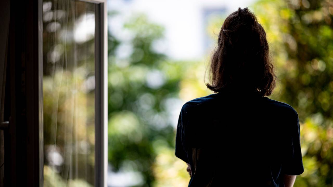 Frau steht mit dem Rücken zur Kamera am Fenster und schaut hinaus.