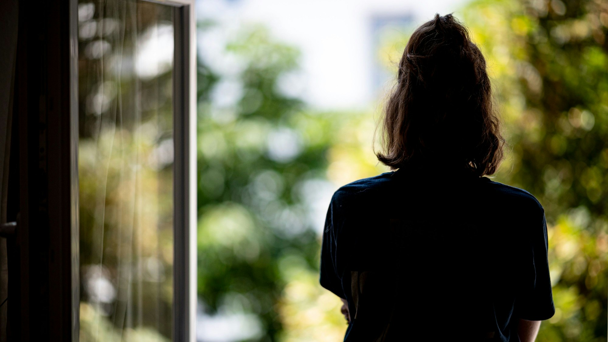 Frau steht mit dem Rücken zur Kamera am Fenster und schaut hinaus.