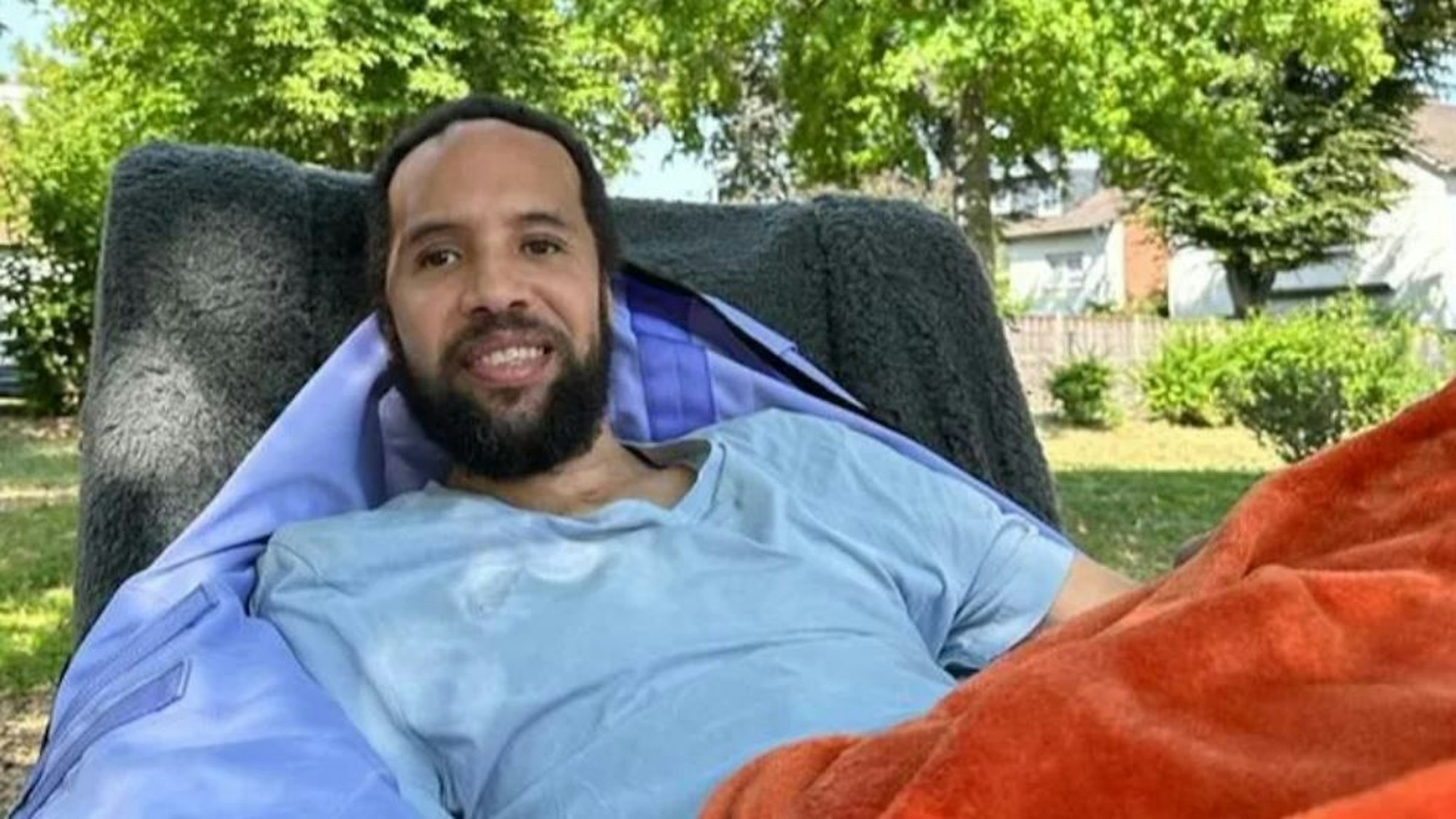 Ein junger Mann in einem blauen Shirt liegt in einem Bett, im Hintergrund sind Bäume zu sehen.