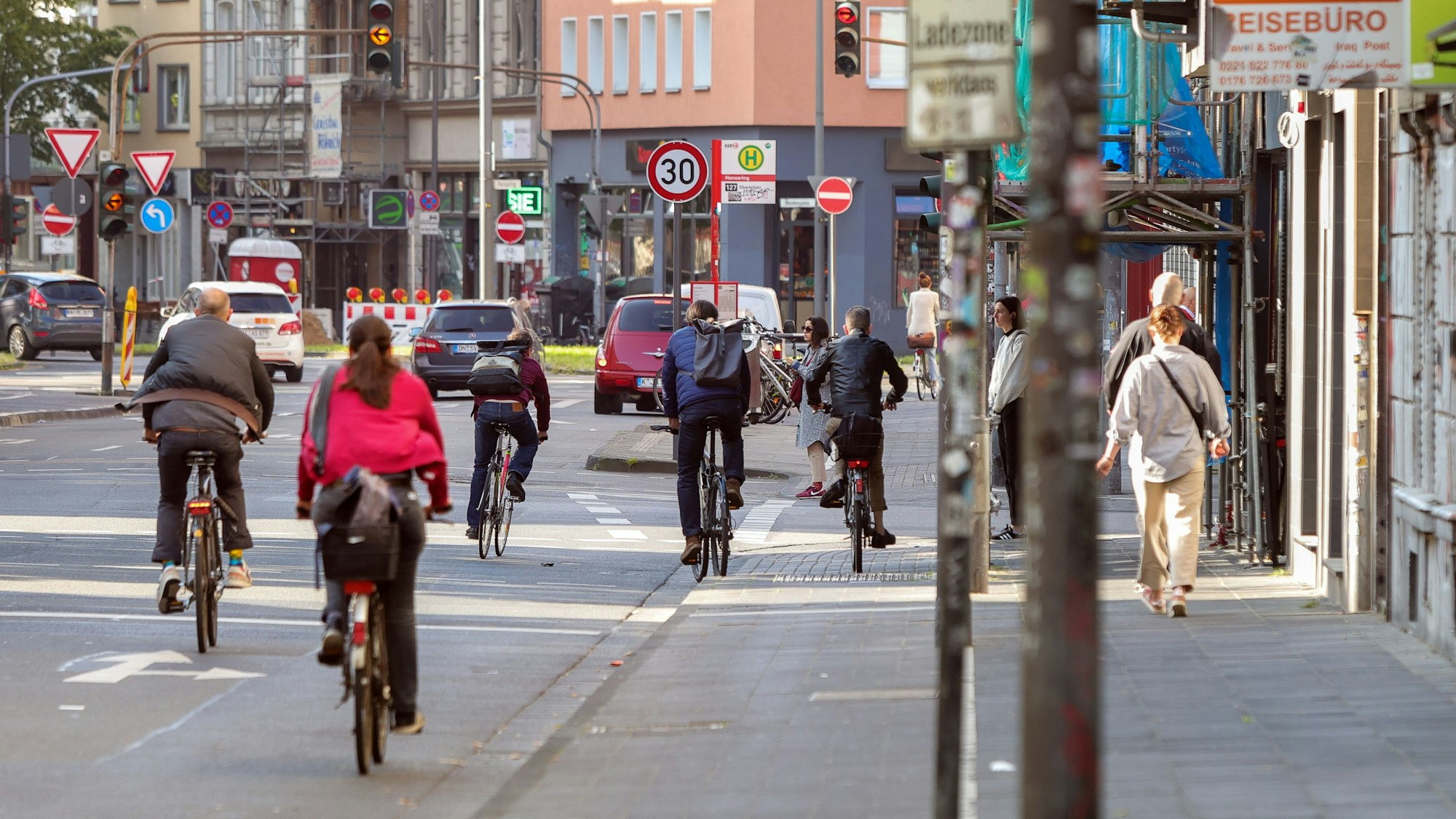 Zahlreiche Menschen sind zu Fuß oder auf dem Fahrrad an einer Straße unterwegs, auch Autos sind zu sehen.