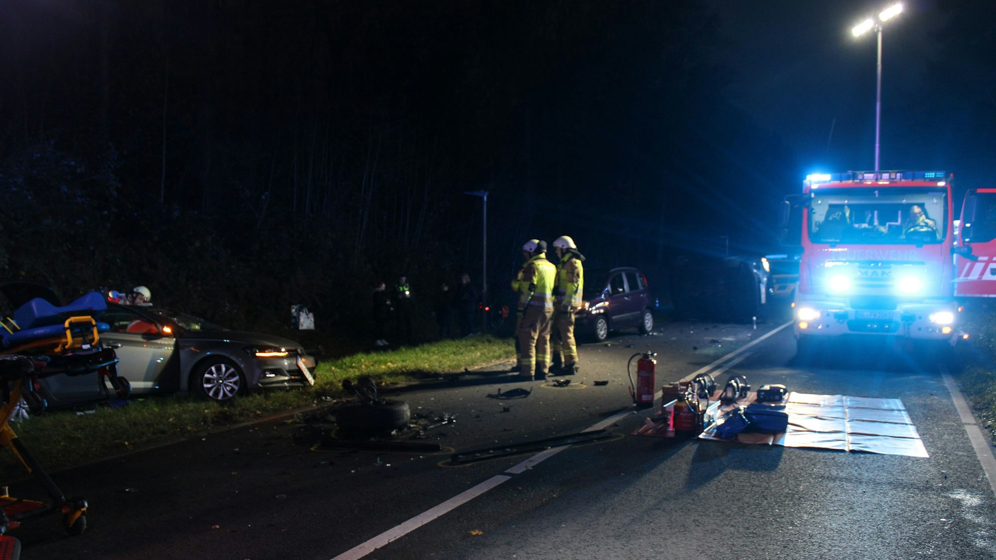 Eine Unfallstelle mit demolierten Autos in der Dunkelheit. Auch ein Feuerwehrauto und Rettungskräfte sind zu sehen.