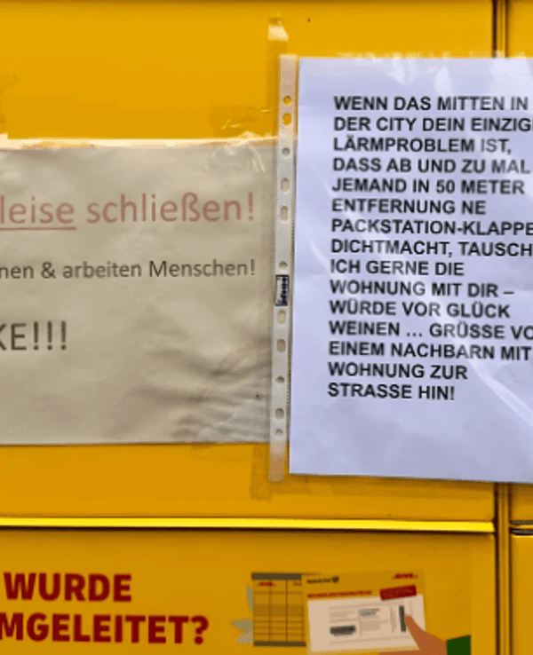 Das Bild zeigt eine DHL-Packstation, an der zwei Zettel hängen, auf denen sich Anwohnenden über den Lärm beschweren