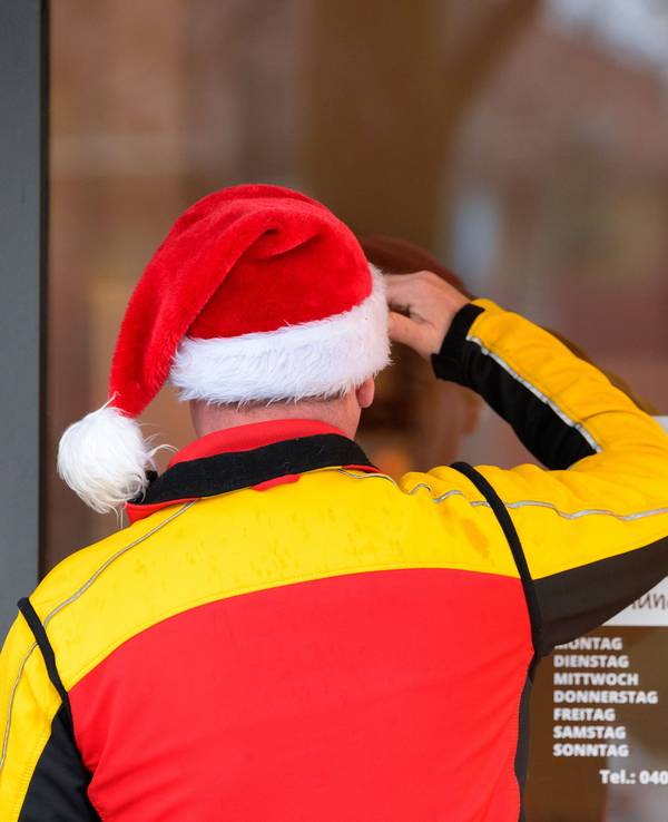 Ein Paketbote des Paketdienstes DHL mit einer Weihnachtsmütze auf dem Kopf schaut suchend durch die Eingangstür eines Ladengeschäfts.