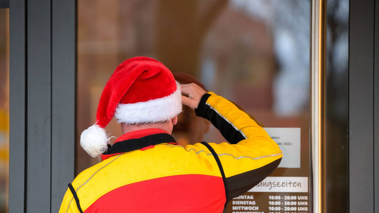 Ein Paketbote des Paketdienstes DHL mit einer Weihnachtsmütze auf dem Kopf schaut suchend durch die Eingangstür eines Ladengeschäfts.
