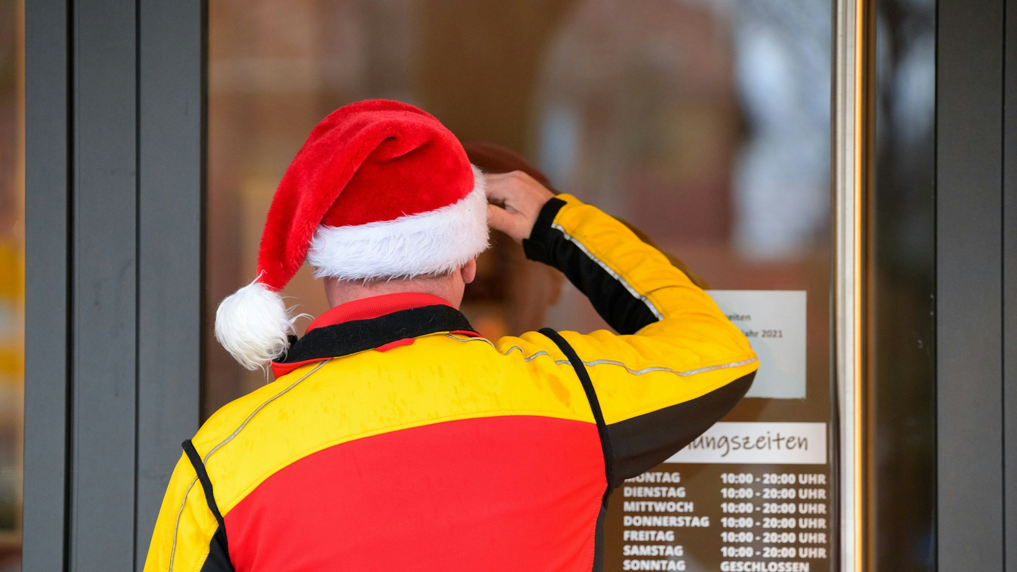 Ein Paketbote des Paketdienstes DHL mit einer Weihnachtsmütze auf dem Kopf schaut suchend durch die Eingangstür eines Ladengeschäfts.