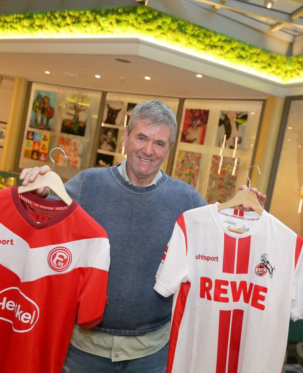 Friedhelm Funkel hält zwei Trikots von Fortuna Düsseldorf und dem 1. FC Köln in die Kamera.