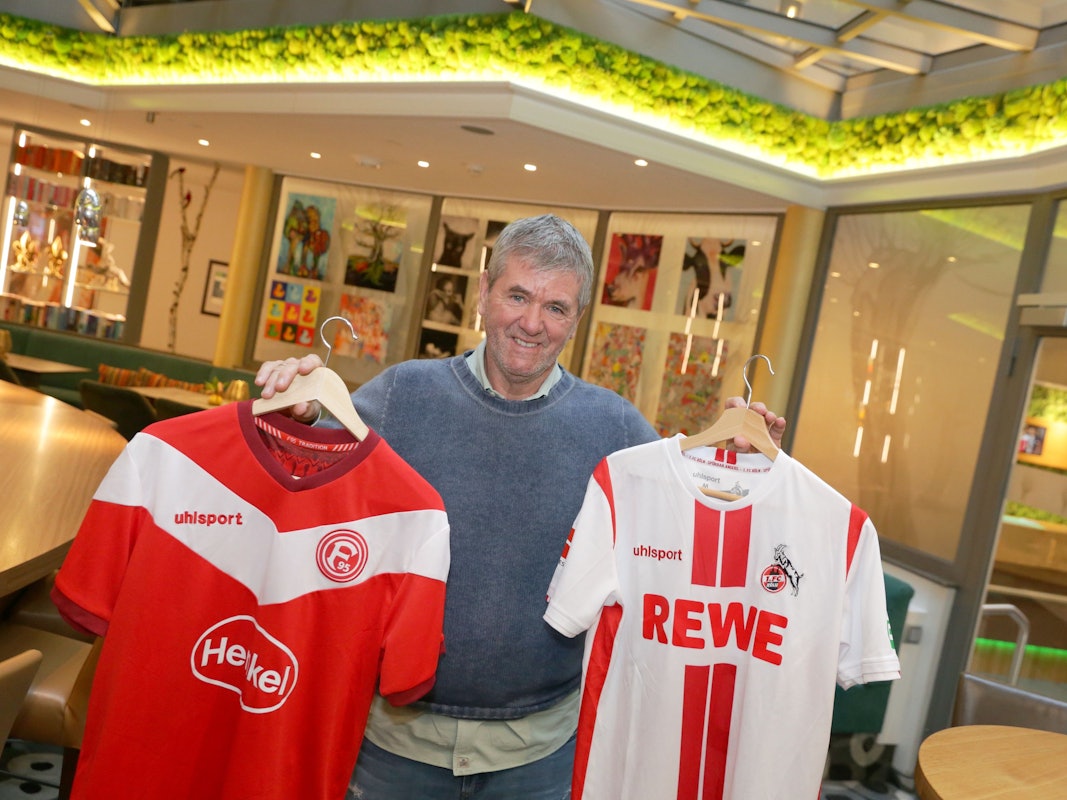 Friedhelm Funkel hält zwei Trikots von Fortuna Düsseldorf und dem 1. FC Köln in die Kamera.