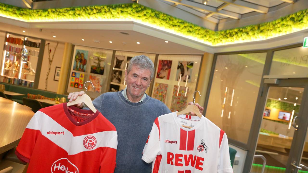 Friedhelm Funkel hält zwei Trikots von Fortuna Düsseldorf und dem 1. FC Köln in die Kamera.