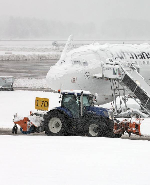 Ein Räum- und Streufahrzeug fährt im Schneetreiben auf dem Flughafen an einer verschneiten Maschine von Lufthansa vorbei.