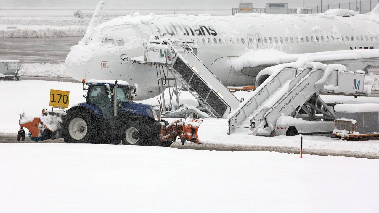 Ein Räum- und Streufahrzeug fährt im Schneetreiben auf dem Flughafen an einer verschneiten Maschine von Lufthansa vorbei.