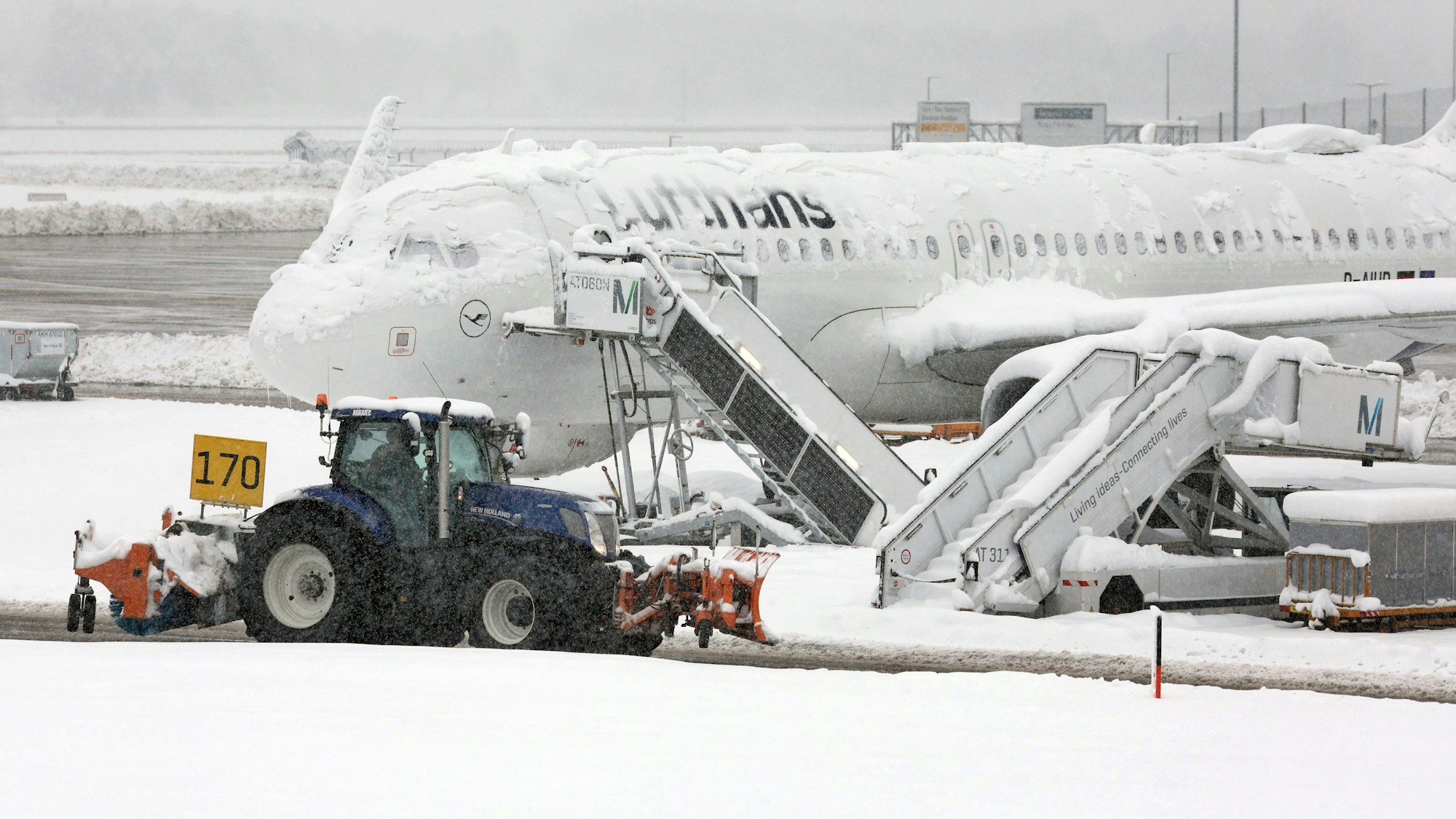 Ein Räum- und Streufahrzeug fährt im Schneetreiben auf dem Flughafen an einer verschneiten Maschine von Lufthansa vorbei.